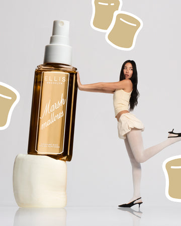 Model with brown hair in yellow tank top and skirt, white tights, and black heels faces to the side, stands on one leg and holds up with her arms a life size bottle of MARSHMALLOWS Perfume Mist without a cap stacked on top of a marshmallow on a gray background with white outlined, beige cartoon marshmallows