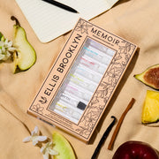 Boîte de découverte de parfums Memoir avec un design floral beige et noir montrant les flacons de 1,5 ml Dear Sky, Miami Nectar, Apple Love, Salt, Sun Fruit, Florist, Myth, Sweet et Vanilla Milk, posés sur un tissu marron clair avec figue, ananas, pomme, bois de santal, gousse de vanille, poire, fleurs blanches et un carnet avec stylo.