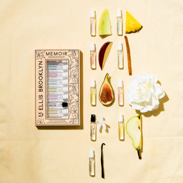 Boîte de découverte de parfums Memoir avec un design floral beige et noir à côté des flacons Dear Sky 1,5 ml, Miami Nectar 1,5 ml, Apple Love 1,5 ml, Salt 1,5 ml, Sun Fruit 1,5 ml, Florist 1,5 ml, Myth 1,5 ml, Sweet 1,5 ml et Vanilla Milk 1,5 ml, sur un fond jaune avec figue, fleur blanche, melon, gousse de vanille, bois de santal, ananas et pomme.
