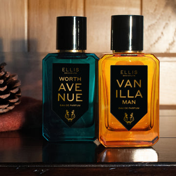 Worth Avenue 50ml Eau De Parfum and Vanilla Man 50ml Eau De Parfum resting on wood shelf next to fabric and pine cone, with wood paneling in background.