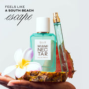 Model hand with light pink nail polish holding Miami Nectar 50ml Eau De Parfum, Miami Nectar 10ml Eau De Parfum Travel Spray without cap, flower, and slice of pink pineapple on large slice of pink pineapple in hand, against white background with black text that reads 