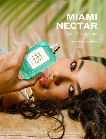 Closeup of green palm leaves and woman with brown hair wearing a gold necklace and blue dress laying on a tan couch and holding MIAMI NECTAR Eau de Parfum 50ml without a cap with white text that reads "MIAMI NECTAR Eau de Parfum starring Jaz Smith"
