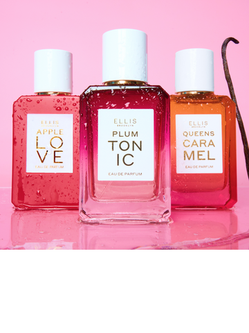 APPLE LOVE Eau de Parfum 50ml, PLUM TONIC Eau de Parfum 50ml, and QUEENS CARAMEL Eau de Parfum 50ml all covered in water droplets next to vanilla bean on top of clear liquid on a pink background