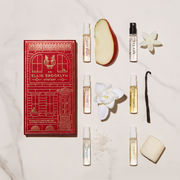 Une boîte rouge Ellis Brooklyn Mystery Fragrance Discovery Set sur une table en marbre blanc avec un flacon Apple Love de 1,5 ml à côté d'une tranche de pomme, un flacon Myth de 1,5 ml à côté d'une fleur blanche, un flacon Florist de 1,5 ml à côté d'une fleur blanche, un flacon Vanilla Milk de 1,5 ml à côté d'une gousse de vanille, un flacon Salt de 1,5 ml à côté de sel marin, et un flacon Sweet de 1,5 ml à côté d'une guimauve.