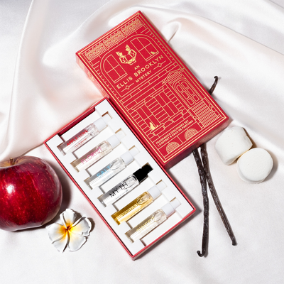 Red An Ellis Brooklyn Mystery Fragrance Discovery Set box opened to show inside and Apple Love 1.5ml Eau De Parfum Vial, Florist 1.5ml Eau De Parfum Vial, Salt 1.5ml Eau De Parfum Vial, Myth 1.5ml Eau De Parfum Vial, Vanilla Milk 1.5ml Eau De Parfum Vial and Sweet 1.5ml Eau De Parfum Vial on white satin sheet with vanilla beans, marshmallows, apple and white flower.