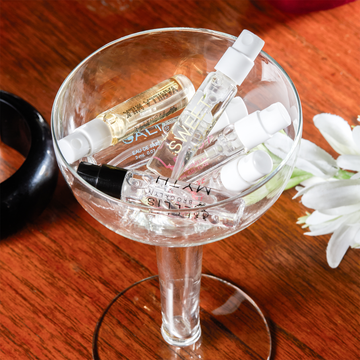 Flacon de parfum Apple Love 1,5 ml, flacon de parfum Florist 1,5 ml, flacon de parfum Salt 1,5 ml, flacon de parfum Myth 1,5 ml, flacon de parfum Vanilla Milk 1,5 ml et flacon de parfum Sweet 1,5 ml dans un verre à pied sur une table en bois à côté de fleurs blanches et d’un bracelet noir.