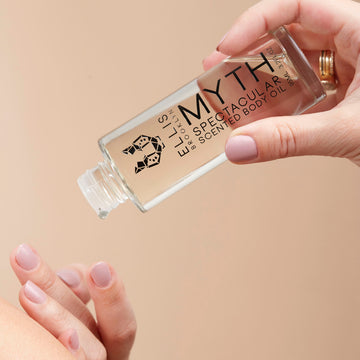 Mannequin avec du vernis à ongles nude portant des bagues en or tenant le Model tenant l'Huile Corporelle Parfumée Spectaculaire Myth 95 ml sans le bouchon dans une main tout en versant le produit sur l'autre main.