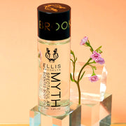 Huile corporelle parfumée Myth Spectacular 95 ml sur un bloc de verre devant des fleurs roses, bloc de verre et cylindre en verre, sur fond orange.