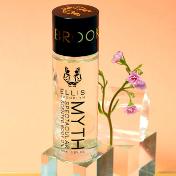 Huile corporelle parfumée Myth Spectacular 95 ml sur un bloc de verre devant des fleurs roses, bloc de verre et cylindre en verre, sur fond orange.