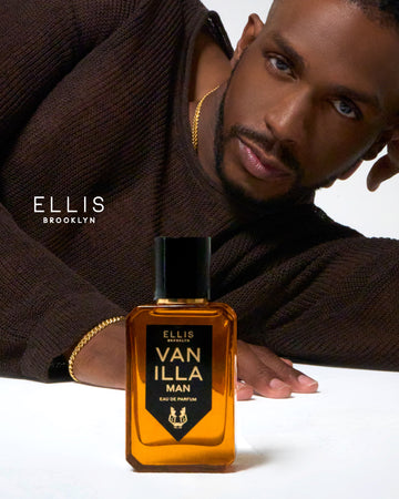 Model with brown hair wearing gold necklaces, bracelets, rings, and brown sweater laying on white ground behind Vanilla Man 50ml Eau De Parfum, and white Ellis Brooklyn logo.