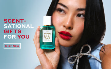 Model with brown hair wearing red lipstick, blue dress, and diamond clip holding Miami Nectar 50ml Eau De Parfum to face, on blue background with red and white text that reads "Scent-Sational Gifts For You" above red text that reads "Shop Now" surrounded by white border.