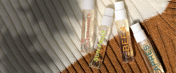 SUPER AMBER Eau de Parfum 1.5ml vial, SUN FRUIT Eau de Parfum 1.5ml vial, BEE Eau de Parfum 1.5ml vial and APRES Eau de Parfum 1.5ml vial laying on white and brown knit fabric with shadows of leaves