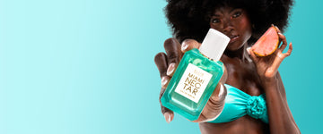 Woman with brown hair wearing a strapless teal bikini top and a French manicure holds MIAMI NECTAR Eau de Parfum 50ml out in front of her in one hand and a slice of pink pineapple in the other hand on a teal and white ombré background