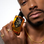 Model wearing gold necklaces, gold rings, and brown sweater holding Vanilla Man 50ml Eau De Parfum to face in one hand, on white background.