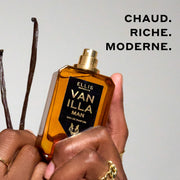 Modèle portant des bagues en or tenant le Vanilla Man 50ml Eau De Parfum avec le bouchon enlevé dans une main, et des gousses de vanille dans l'autre, sur un fond gris clair avec un texte noir qui dit « Chaud. Riche. Moderne. »  french_image