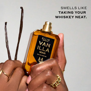 Model weaing gold rings holding Vanilla Man 50ml Eau De Parfum with cap removed in one hand, and vanilla beans in the other, on light grey background with black text that reads "Smells like taking your whiskey neat."