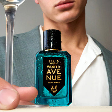 Modèle portant une veste grise et une chemise blanche, tenant la tige d'un verre à côté du parfum Worth Avenue 50ml Eau De Parfum couvert de gouttelettes d'eau sur une table blanche, sur fond gris clair.