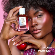 Woman with brown hair wearing a purple sequin tank top and pink highlighter holds PLUM TONIC Eau de Parfum 50ml against her head in one hand and a plum in the other hand on a pink background with white text that reads 