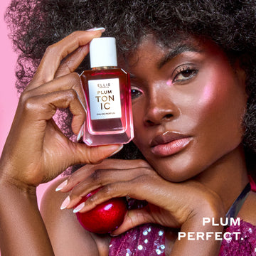 Woman with brown hair wearing a purple sequin tank top and pink highlighter holds PLUM TONIC Eau de Parfum 50ml against her head in one hand and a plum in the other hand on a pink background with white text that reads 