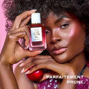 Une femme aux cheveux bruns portant un débardeur à sequins violet et du surligneur rose tient PLUM TONIC Eau de Parfum 50 ml contre sa tête d'une main et une prune de l'autre sur un fond rose avec un texte blanc qui dit « Parfaitement prune. »