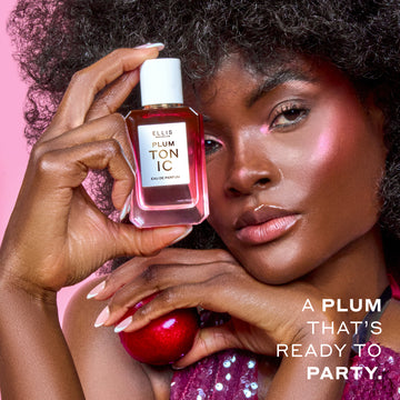 Woman with brown hair wearing a purple sequin tank top and pink highlighter holds PLUM TONIC Eau de Parfum 50ml against her head in one hand and a plum in the other hand on a pink background with white text that reads "A plum that's ready to party."