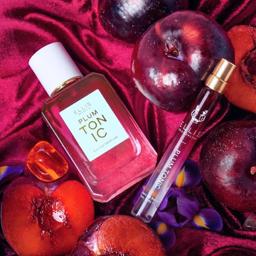 PLUM TONIC Eau de Parfum 50ml and PLUM TONIC Eau de Parfum 10ml next to whole plums, sliced plums, amber stones, and purple irises on red velvet fabric