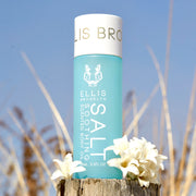 Huile corporelle parfumée apaisante au sel 95ml à côté de fleurs blanches sur un morceau de bois avec des dunes de plage et le ciel en arrière-plan.