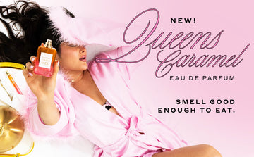 Model with black hair and French manicure wearing gold necklace and pink satin robe with feathers at cuffs laying down on pink couch next to gold tray with napkin, fork, and Queens Caramel 10ml Eau De Parfum Travel Spray, holding Queens Caramel 50ml Eau De Parfum without cap to camera on pink background with text that reads "New! Queens Caramel Eau De Parfum Smell Good Enough To Eat."