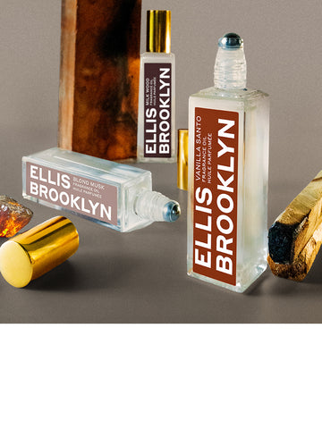 Vanilla Santo 15ml standing on end with cap removed and piece of burnt wood, next Blond Musk 15ml laying on side with cap removed and piece of amber, in front of Milk Wood 15ml standing on end next to piece of wood, on grey background.