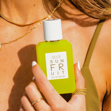 Closeup of woman with brown hair and a white manicure wearing a green bikini top, a gold link necklace, and gold rings holds SUN FRUIT Eau de Parfum 50ml against her chest
