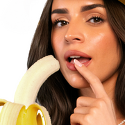 Model with brown hair and finger placed against her open lips, looking into camera while holding a peeled banana with a bite taken out of it on a white background