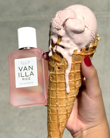 Hand with red manicure holds VANILLA RICE Eau de Parfum 50ml and an ice cream cone with pink ice cream on gray concrete background