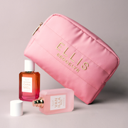 Pink Cosmetic Bag with gold embroidery that reads "Ellis Brooklyn" propped up on QUEENS CARAMEL Eau de Parfum 50ml and behind VANILLA RICE Eau de Parfum 50ml without cap laying down next to cap on gray background