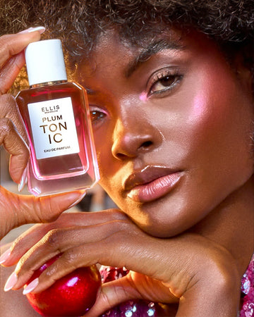 Woman with brown hair wearing a purple sequin tank top and pink highlighter holds PLUM TONIC Eau de Parfum 50ml against her head in one hand and a plum in the other hand