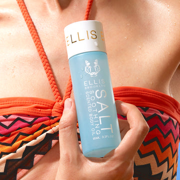 Mannequin portant un haut de bikini orange, rose et marron, tenant l'huile corporelle parfumée apaisante Salt 95ml d'une main près de la poitrine, sur fond bleu.