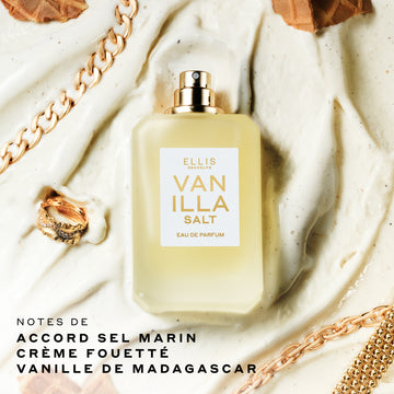 Bouteille de 50 mL de parfum Vanille Sel présentée sur un fond couleur crème avec des bijoux en or et des morceaux de gaufre, avec un texte noir en bas à gauche indiquant « notes de accord sel marin, crème fouettée, vanille de Madagascar ».