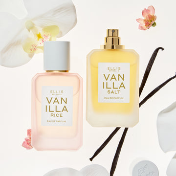 Vanilla Rice and Vanilla Salt Eau de Parfum 50mL bottles surrounded by white orchids, pink flowers, and vanilla beans on a white background. Vanilla Rice is shown in a soft pink bottle with a white cap, and Vanilla Salt in a golden yellow bottle with a gold spray top.