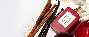 APPLE LOVE Eau de Parfum 50ml without cap lays down surrounded by two apples, two vanilla beans, 5 sticks of wood, sugar crystals, a white cap and a white flower all on a white background