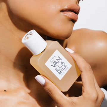 Modèle avec du vernis à ongles blanc et un piercing en diamant au-dessus de la lèvre tenant Vanilla Milk 50ml Eau De Parfum près de la mâchoire, sur fond blanc.