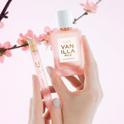 Des mains arborant une manucure rose pâle tiennent l'eau de parfum VANILLA RICE (50 ml) et l'eau de parfum VANILLA RICE (10 ml) sur un fond rose agrémenté de fleurs de cerisier (sakura) roses.