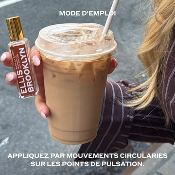 Mannequin aux ongles nus et cheveux blonds portant une chemise à manches longues rayée brun et blanc, tenant un café glacé et une huile parfumée Vanilla Santo 15 ml dans une main, dans une rue de la ville, entre du texte blanc qui dit « Appliquez par mouvements circularies sur les points de pulsation ».