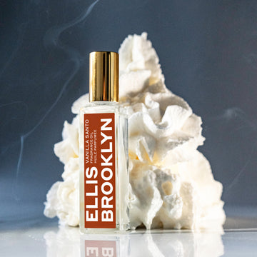 Huile parfumée Vanille Santo 15 ml devant un tourbillon de crème fouettée sur une table en verre avec un fond gris et de la fumée.