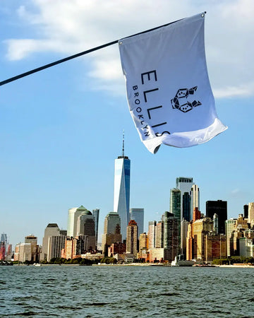 white flag with Ellis Brooklyn Logo on pole over water, against sky and clouds, with New York City buildings in background.