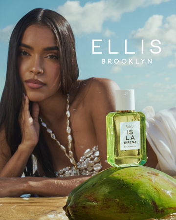 Woman with brown hair wearing a white shell halter neck bikini top and white skirt lays down on her side in wet sand with ISLA SIRENA Eau de Parfum 50ml on a green coconut in front of her with white text that reads 'Ellis Brooklyn' and a blue sky and white clouds in the background