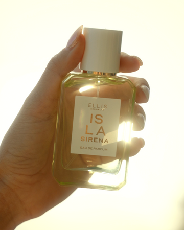 Hand with light pink manicure holds ISLA SIRENA Eau de Parfum up against the light with light shining through the bottle against a white background