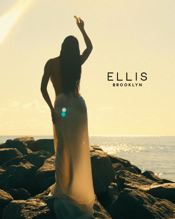 Woman with wet brown hair wearing a white shell bikini top and white skirt faces away while standing on a jetty of rocks and holding ISLA SIRENA Eau de Parfum in her hand above her head with black text that reads 'Ellis Brooklyn' and an ocean and yellow sunlit sky in the background