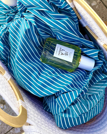 White rope bag with light wooden handles sits on tan cement sidewalk with a blue and white striped shirt and ISLA SIRENA Eau de Parfum 50ml laying on its side inside
