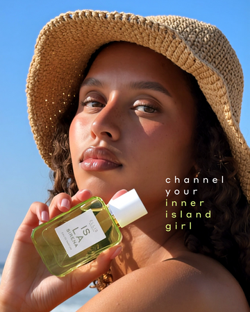 Woman with brown, wavy hair wearing a tan woven hat looks over her shoulder and holds ISLA SIRENA Eau de Parfum 50ml against her chin with white and green text that reads 'channel your inner island girl' on a blue background