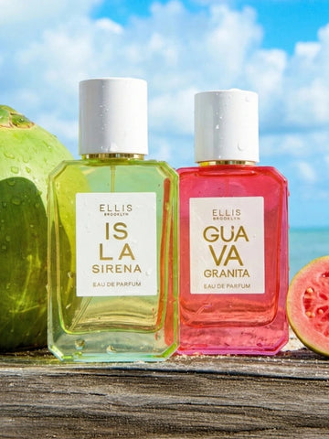 ISLA SIRENA Eau de Parfum 50ml sits next to GUAVA GRANITA Eau de Parfum 50ml between a green coconut and half of a sliced guava all covered in water droplets on top of brown wood with a blue sky and white clouds in the background