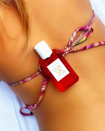 Model with blonde hair wearing multi colored bikini top laying on white towel with Guava Granita 50ml Eau De Parfum on back.